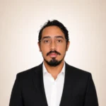 Man with glasses and a beard wearing a black suit and white shirt, facing forward against a plain background.