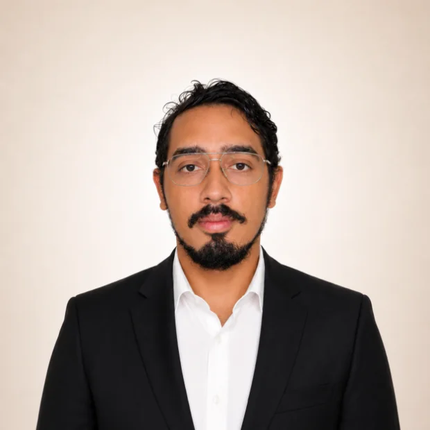 Man with glasses and a beard wearing a black suit and white shirt, facing forward against a plain background.