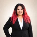 Woman with wavy red hair in a black blazer and white shirt, standing and smiling at the camera.