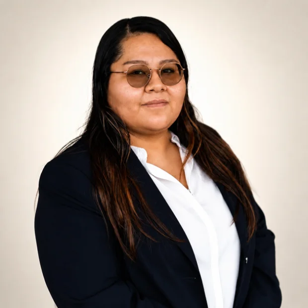 Person with long dark hair, glasses, and a navy blazer over a white shirt, posing against a plain background.