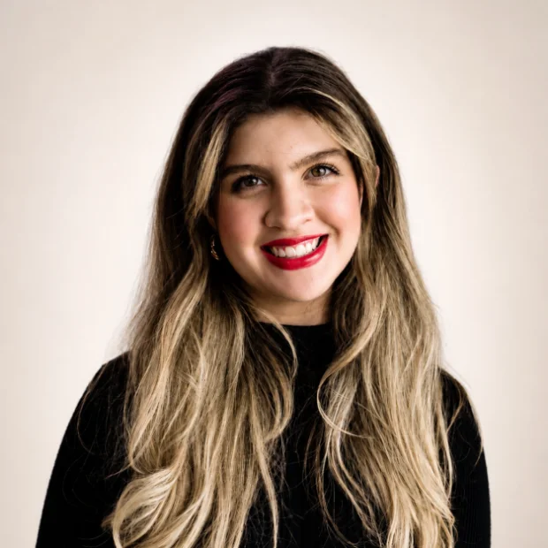 Smiling woman with long, wavy blonde hair wearing a black top stands in front of a plain background.